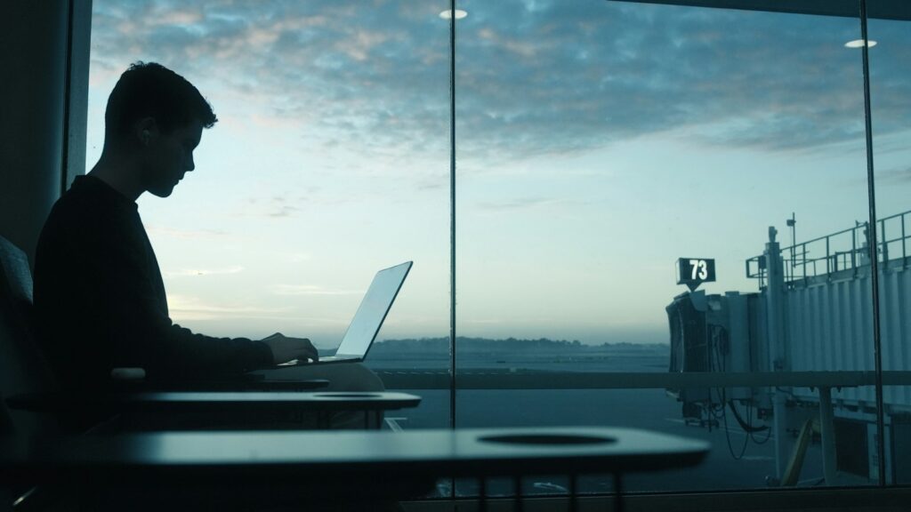 Mann arbeitet bei Sonnenaufgang am Laptop vor einem Flughafenfenster – digitale Mobilität im Geschäftsalltag